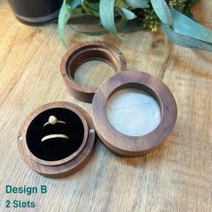 K&ouml;nnte beinhalten: Eine Holzkiste f&uuml;r Ringe mit zwei F&auml;chern, eines mit einem Ring darin. Die Kiste ist aus dunklem Holz und hat ein kreisf&ouml;rmiges Design. Der Text "Design B 2 Slots" ist unterhalb der Kiste sichtbar.