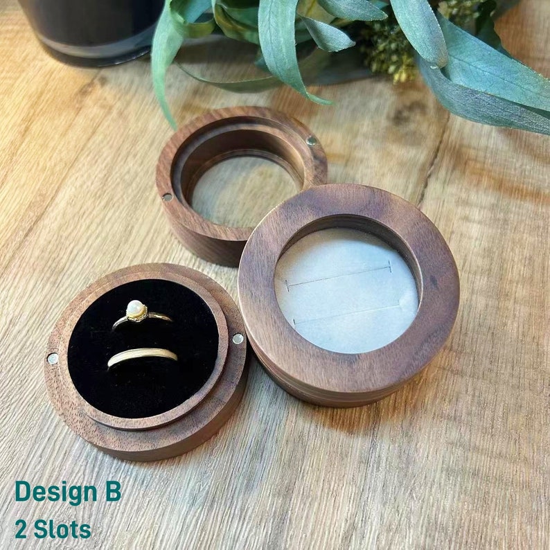 K&ouml;nnte beinhalten: Eine Holzkiste f&uuml;r Ringe mit zwei F&auml;chern, eines mit einem Ring darin. Die Kiste ist aus dunklem Holz und hat ein kreisf&ouml;rmiges Design. Der Text "Design B 2 Slots" ist unterhalb der Kiste sichtbar.