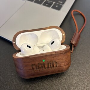 May include: A wooden case for wireless earbuds with a personalized inscription that reads "DAVID". The case is brown with a light brown leather strap.