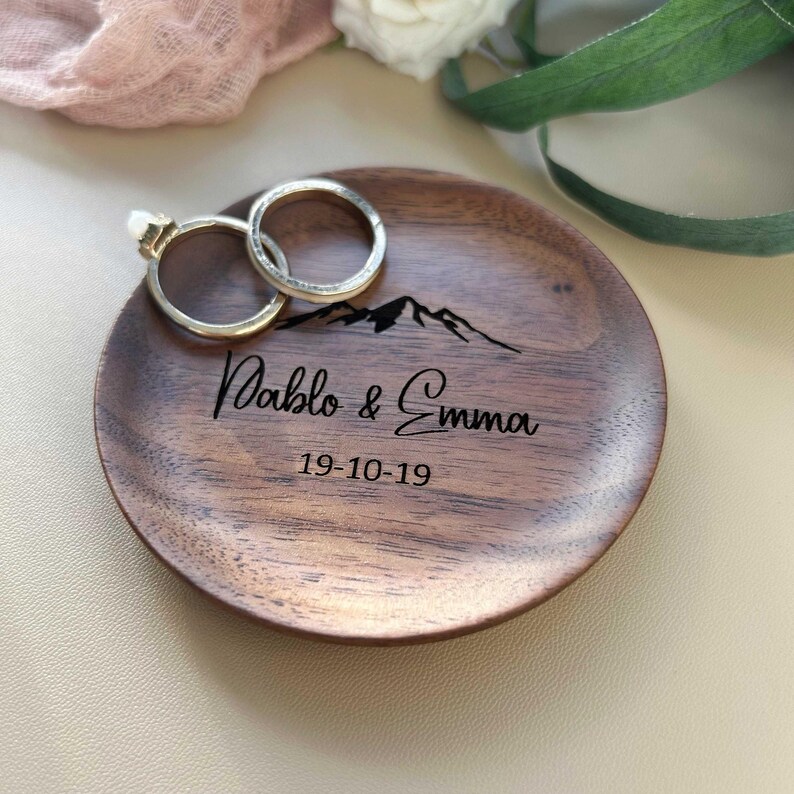 Może przedstawiać: Drewniana podstawka na obrączki z wzorem sylwetki g&oacute;r, spersonalizowana z imionami "Pablo & Emma" i datą "19-10-19". Na podstawce leżą dwa złote obrączki.