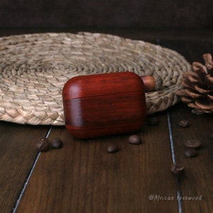 May include: A dark red wooden case for wireless earbuds. The case has a smooth, polished finish and a small, round knob on the top. The case is sitting on a wooden surface with coffee beans scattered around it.