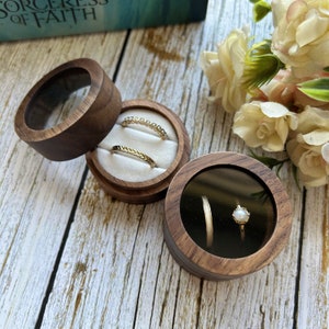 Pode incluir: Três caixas de anéis de madeira com tampas transparentes, uma aberta e duas fechadas, exibindo anéis de ouro. As caixas são redondas com acabamento em madeira escura e interior branco. Um buquê de flores está ao fundo.