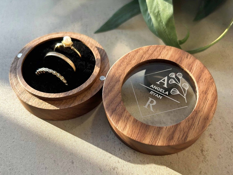 K&ouml;nnte beinhalten: Eine Holzkiste f&uuml;r Ringe mit einem durchsichtigen Deckel. Der Deckel ist mit den Namen "Angela Ryan" und den Initialen "A" und "R" graviert. Die Kiste ist ge&ouml;ffnet und enth&auml;lt drei Ringe.