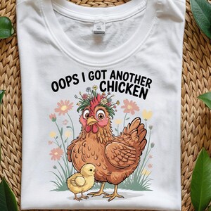 Peut inclure: T-shirt blanc avec une illustration de dessin animé d'une poule portant une couronne de fleurs et tenant un poussin. Le texte "Oops I got another chicken" est imprimé sur le t-shirt.