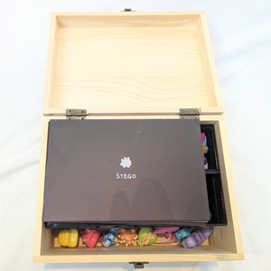 May include: A wooden box with a hinged lid, open to reveal a black insert with the word "STEGO" printed on it. The box is filled with colourful figurines.