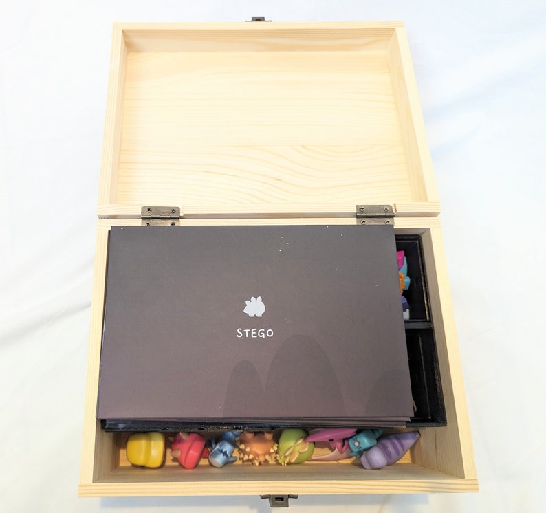 May include: A wooden box with a hinged lid, open to reveal a black insert with the word "STEGO" printed on it. The box is filled with colourful figurines.