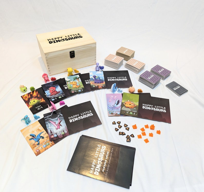 May include: A wooden box with the text "Happy Little Dinosaurs" on the lid. The box is open and contains a variety of game pieces, including small dinosaur figurines, cards, and dice. The game pieces are arranged on a white surface.