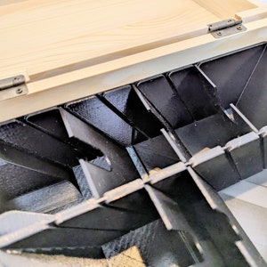 May include: A wooden box with a hinged lid, revealing a black plastic insert with multiple compartments. The compartments are angled and designed to hold small items.