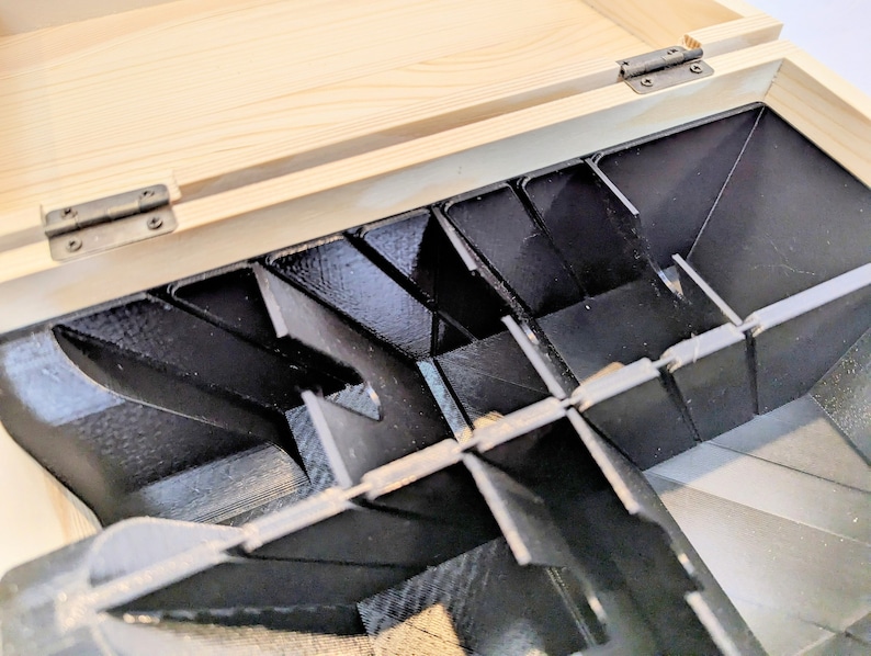 May include: A wooden box with a hinged lid, revealing a black plastic insert with multiple compartments. The compartments are angled and designed to hold small items.