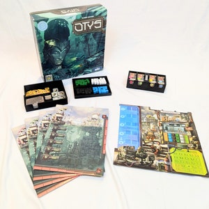 以下が含まれることがあります： 「Otys」というボードゲーム。緑と青の箱、ゲームボード、さまざまなゲームピースが入っています。ゲームボードは、建物や通りがある街の風景です。ゲームピースには、サイコロ、トークン、カードが含まれています。