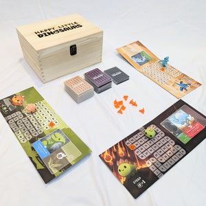May include: A wooden box with the text "Happy Little Dinosaurs" on the lid. The box contains a board game with colourful game boards, cards, and dinosaur figurines.