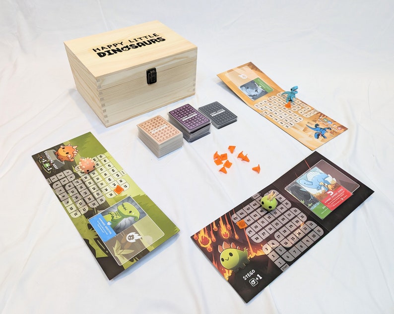 May include: A wooden box with the text "Happy Little Dinosaurs" on the lid. The box contains a board game with colourful game boards, cards, and dinosaur figurines.