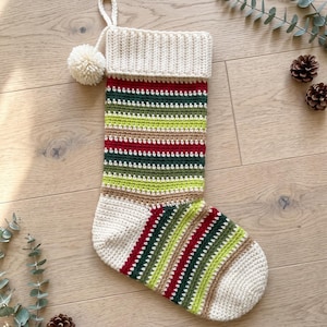 Puede incluir: Un calcetín navideño de ganchillo con rayas horizontales en rojo, verde oscuro, verde lima y beige. El calcetín tiene un puño de color crema sólido, talón y puntera a juego, y un pompón de color crema unido a un lazo para colgar.
