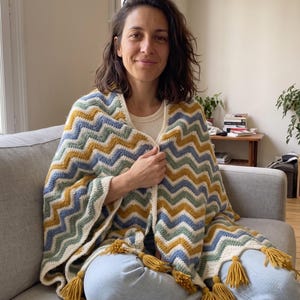 May include: A crochet shawl featuring a chevron pattern in mustard yellow, sage green, light blue, and cream. The edges are finished with a cream border and mustard yellow tassels along the bottom hem, creating a textured, layered appearance.