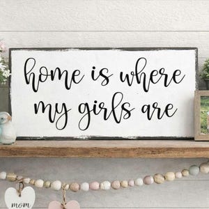 May include: White wooden sign with black text that reads "home is where my girls are". The sign is distressed and has a black border.