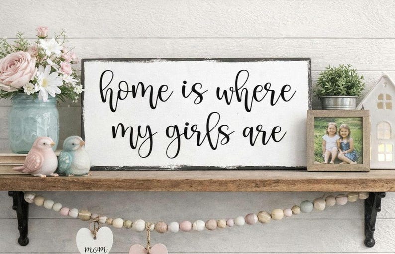 May include: White wooden sign with black text that reads "home is where my girls are". The sign is distressed and has a black border.