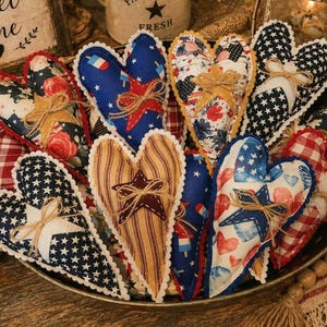 May include: A collection of patriotic-themed heart-shaped fabric ornaments. Each heart features unique patterns, including stars, stripes, and floral designs, with decorative twine bows. The ornaments are arranged in a metal tray, evoking a rustic, Americana aesthetic.
