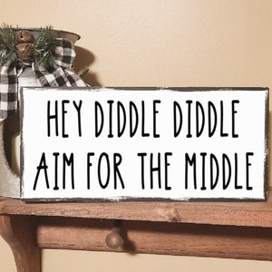 May include: A white wooden sign with black text that reads "Hey Diddle Diddle Aim For The Middle". The sign is sitting on a wooden shelf.