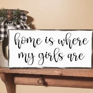 May include: White wooden sign with black text that reads "home is where my girls are". The sign is distressed and has a black border.