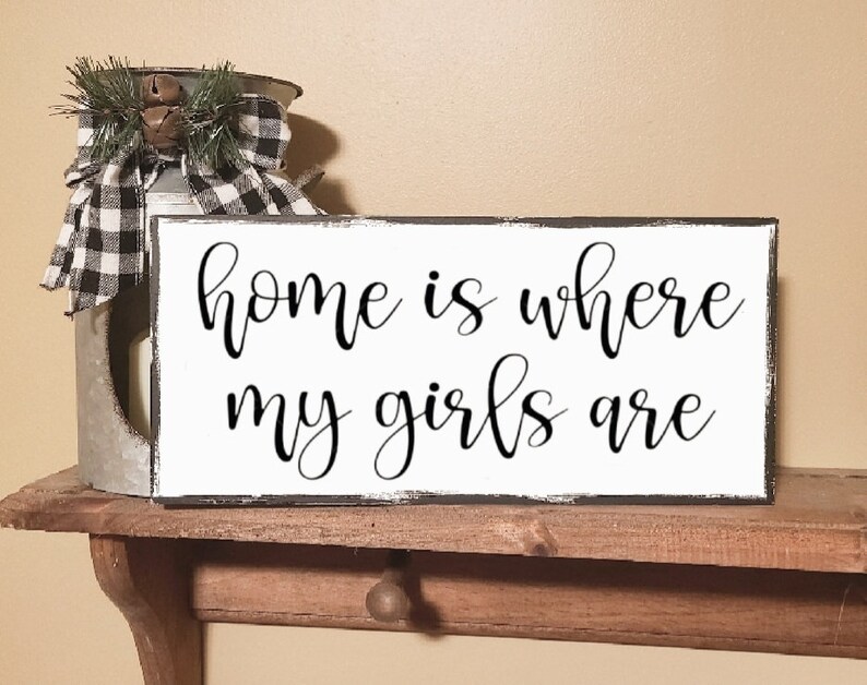 May include: White wooden sign with black text that reads "home is where my girls are". The sign is distressed and has a black border.