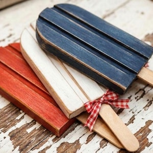 May include: Three wooden popsicle-shaped decorations in red, white, and blue. Each has a wooden stick and the white one is adorned with a red and white checkered bow. The items are arranged on a distressed, white-painted wooden surface.