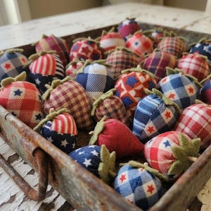 May include: A metal tray filled with fabric strawberries in various patriotic patterns. The strawberries feature red, white, and blue fabrics with stars and stripes, and green felt leaves. The tray has a rustic, weathered appearance.