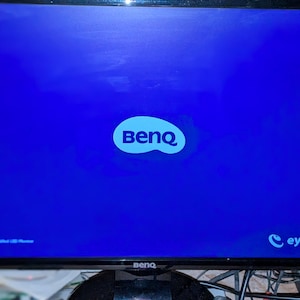 May include: A black BenQ computer monitor displaying a bright blue screen with the BenQ logo in white. The words "eye-care" are visible in the lower right corner. The monitor is on a wooden surface.