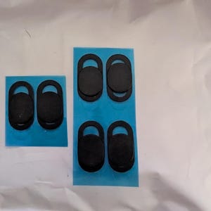 May include: Six black oval webcam privacy covers arranged on two rectangular blue adhesive backing sheets. Each cover features a sliding mechanism to open and close the lens aperture, designed for attachment to electronic device screens.