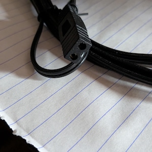 May include: A black power cord with a two-prong connector. The cord is coiled and lies on a white paper with blue lines. A red-tipped component is visible at the end of the cord.
