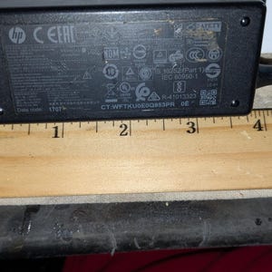 May include: A black HP power adapter with various symbols and text, including "CEEAC" and "IEC 60950-1." A wooden ruler, marked in inches, is positioned below the adapter. The adapter measures approximately 3 inches in length.