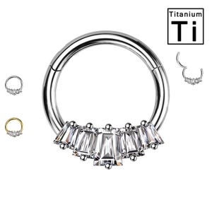 May include: Titanium body jewelry featuring a circular hoop with a row of clear baguette-shaped gems. The hoop is made of titanium and is marked with the symbol "Ti".