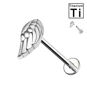 Puede incluir: Un labret de titanio plateado con un diseño en forma de ala. El ala tiene una estructura esquelética detallada. El labret presenta una parte trasera plana en forma de disco y un poste recto. La palabra "Titanium" y el símbolo "Ti" están en la esquina superior derecha.