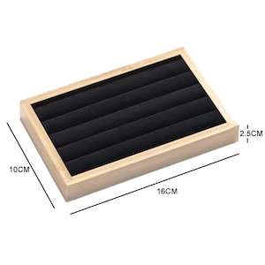Könnte beinhalten: Ein rechteckiges Schmucktablett aus Holz mit schwarzem Samtinnenraum. Das Tablett ist 16 cm lang, 10 cm breit und 2,5 cm hoch. Es hat mehrere Schlitze zum Ordnen von Ringen und anderen kleinen Schmuckstücken.