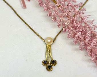 May include: A gold necklace with a pearl pendant and three blue gemstone accents. The necklace is on a white background with pink flowers.