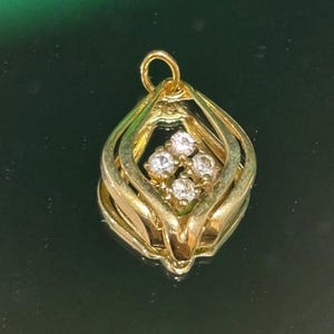 Könnte beinhalten: Ein goldener Anhänger mit einem rautenförmigen Rahmen aus geschwungenen, geschichteten Metallkanten. In der Mitte sind vier kleine, klare, rund geschliffene Steine in einem quadratischen Cluster angeordnet. Oben befindet sich eine kleine Öse.