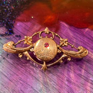 Op de afbeelding: Een goudkleurige metalen broche met een langwerpig, sierlijk frame met kruldetails en kleine bloemaccenten. In het midden toont een ronde schijf gegraveerde lijnen en een enkele, kleine, ronde, felroze edelsteen in het midden geplaatst.