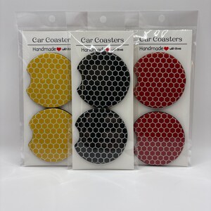 May include: Three sets of car coasters in clear plastic packaging. Each set contains two coasters with a honeycomb pattern. Colors include yellow, black, and red. The packaging reads "Car Coasters Handmade with love."