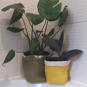 May include: Two potted plants with green leaves. The larger plant is in a brown burlap pot with a white fabric liner. The smaller plant is in a yellow burlap pot with a white fabric liner.