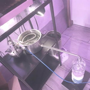 Pode incluir: Um aparelho de destilação com panelas, tubos e uma garrafa de vidro de aço inoxidável. A configuração está em uma prateleira de metal preta com uma luz e um aparelho rosa no fundo. Um tubo azul conecta os componentes.