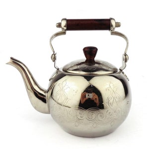 Könnte beinhalten: Eine silberne Teekanne mit einem Holzgriff und einem verzierten Design. Die Teekanne hat einen runden Korpus und einen Ausguss an der Seite.