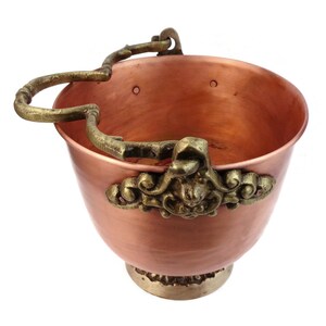 May include: A copper bucket with a decorative brass handle and ornate detailing. The bucket has a rounded shape with a brass base and decorative accents. The handle is curved and attached to the rim.