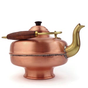 May include: A copper teapot with a wooden handle and a brass spout. The teapot has a rounded body and a small, round base.
