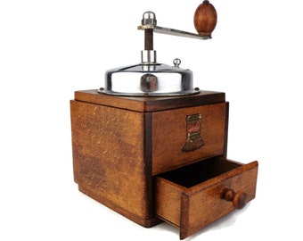 May include: A vintage wooden coffee grinder with a metal crank and a drawer for storing coffee beans. The grinder is made of dark wood with a light wood drawer.