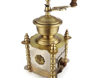 May include: A vintage brass coffee grinder with a wooden handle. The grinder has a square base with decorative details and a round top. The grinder is made of brass and has a silver-colored metal panel on the front.