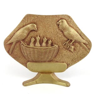 May include: A gold-colored, decorative plaque featuring a bird family. Two adult birds flank a woven basket with four chicks. The plaque is mounted on a small, gold-colored stand, creating a three-dimensional effect. The background has a textured appearance.