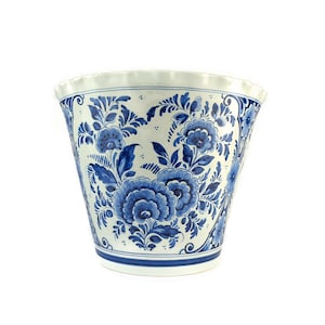 Può includere: Un vaso di ceramica bianca con un motivo floreale blu e bianco. Il vaso ha una base larga e arrotondata e un bordo svasato.