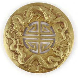 Peut inclure: Une pièce décorative circulaire de couleur dorée, ornée de quatre dragons détaillés entourant un motif géométrique central. Les dragons sont sculptés avec précision et la pièce a un aspect légèrement vieilli.
