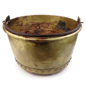 Pode incluir: Um balde de latão vintage com um corpo arredondado e uma borda escura. O balde tem duas pequenas alças e é rebitado ao longo da parte inferior. O interior mostra sinais de idade e uso, com descoloração escura.
