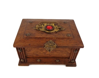 May include: A wooden box with a red jewel and ornate brass accents. The box has a hinged lid and is decorated with metal studs.