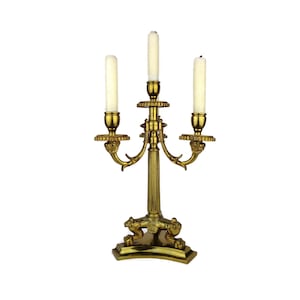 Può includere: Un candelabro in metallo dorato con tre portacandele. Il candelabro ha una base decorativa con un motivo a rotolo e una colonna centrale con dettagli decorativi. I portacandele sono adornati con accenti decorativi.